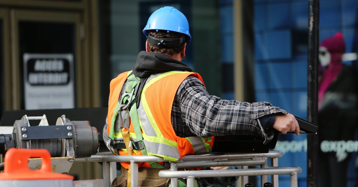 A construction worker wearing a blue helmet and safety vest operates equipment at an urban construction site.