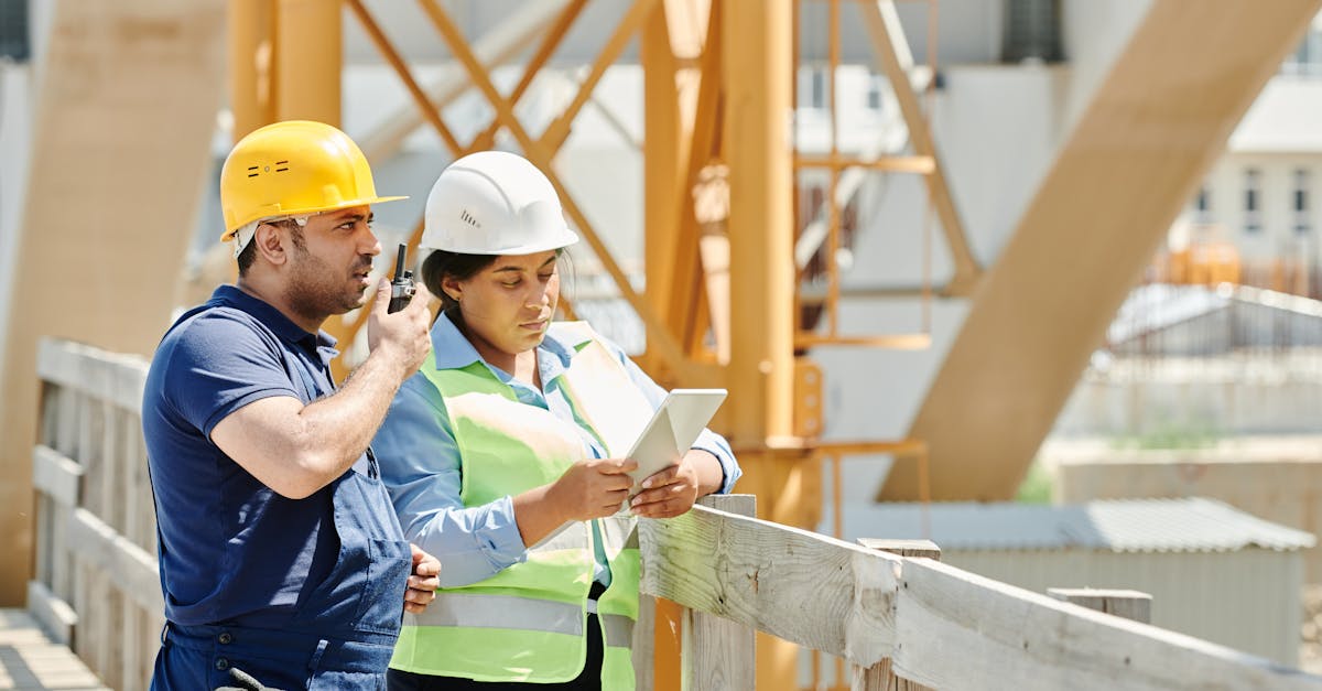 Two construction workers in hardhats discuss plans using a tablet and walkie talkie on-site.